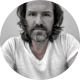 Black-and-white close-up portrait of a man with long hair and a full beard, wearing a white shirt and looking at the camera.